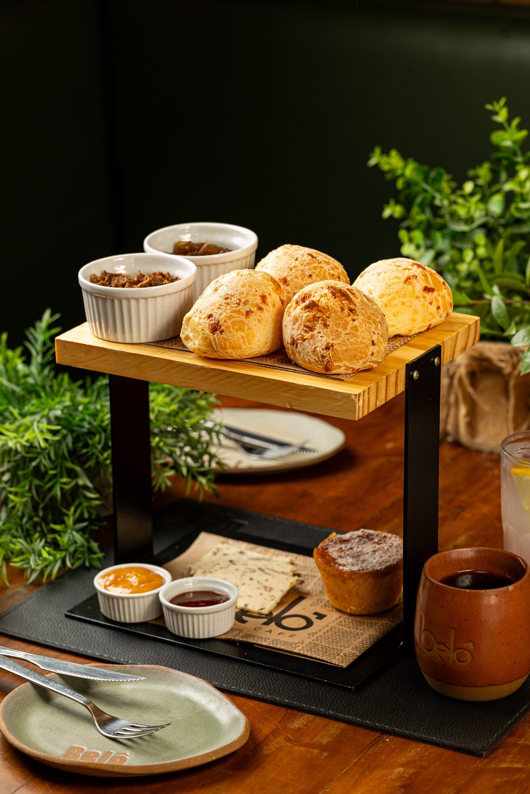 Suporte de dois andares com pães de queijo no topo e, embaixo, tortinha, geleias, biscoito com o selo Belô e um copo de café, sobre tábua de ardósia.