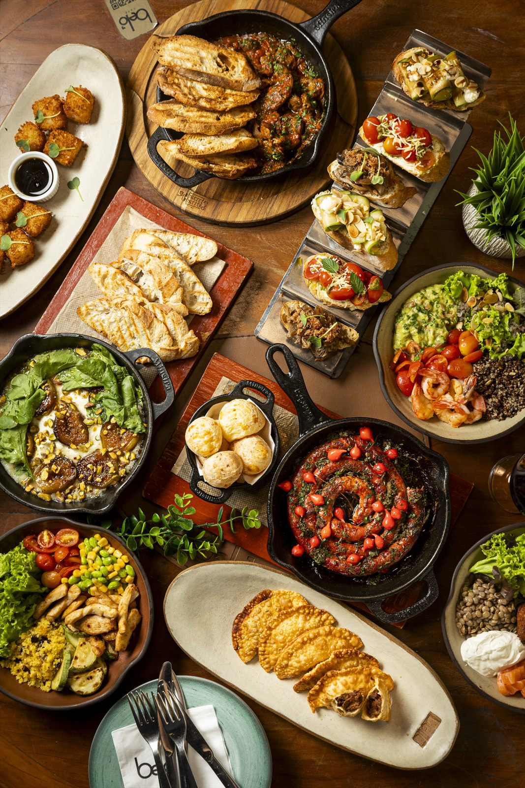 Mesa farta vista de cima com frigideiras de ferro: bruschettas, pão de queijo, ovos, carnes, bolinhos e folhas verdes.