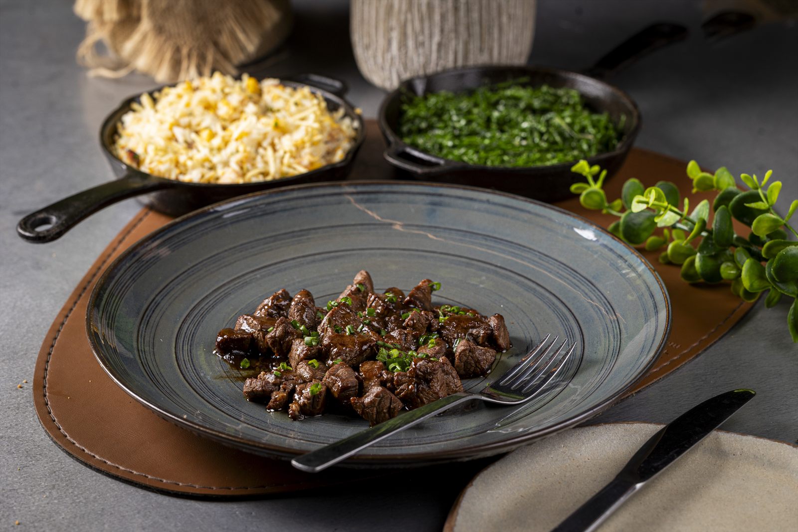 Cubos de carne ao molho com cebolinha em prato esmaltado, arroz e couve em frigideiras de ferro ao fundo.