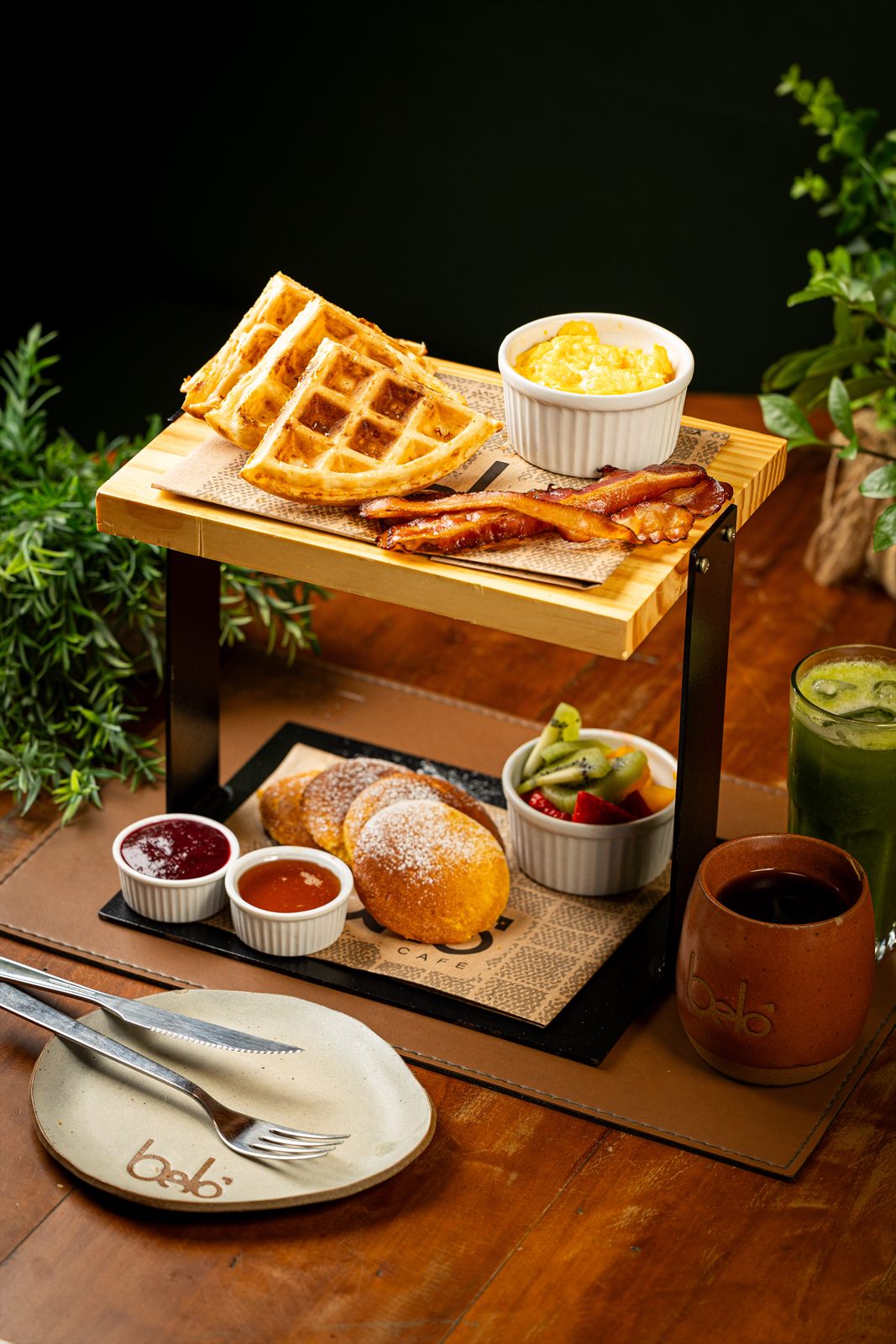 Suporte de dois andares com waffles, bacon e creme em cima; pão doce, geleias e frutas embaixo, acompanhados de suco verde e café.