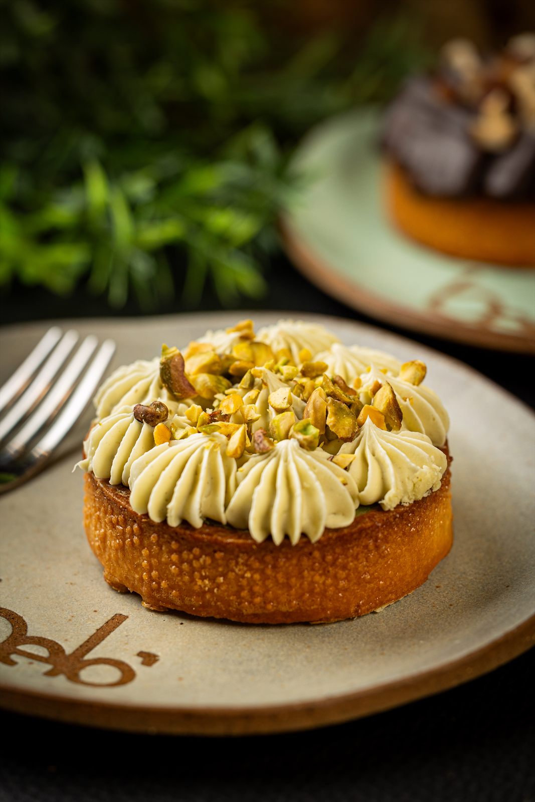 Torta individual com creme de pistache em rosetas e pistaches picados, em prato de cerâmica com selo da casa.