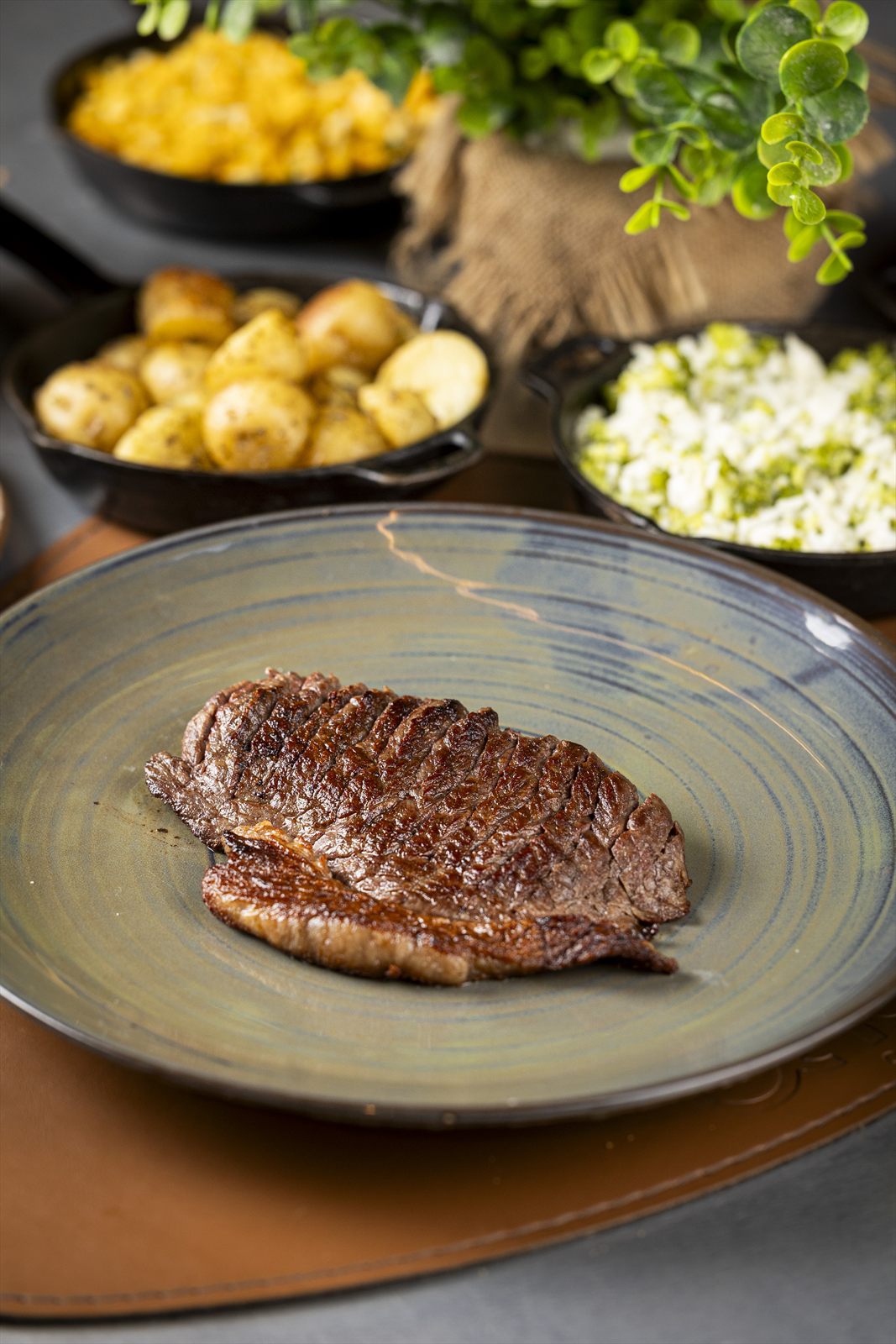 Bife grelhado em prato cinza com batatas douradas, farofa e arroz à brasileira em frigideiras de ferro ao fundo.