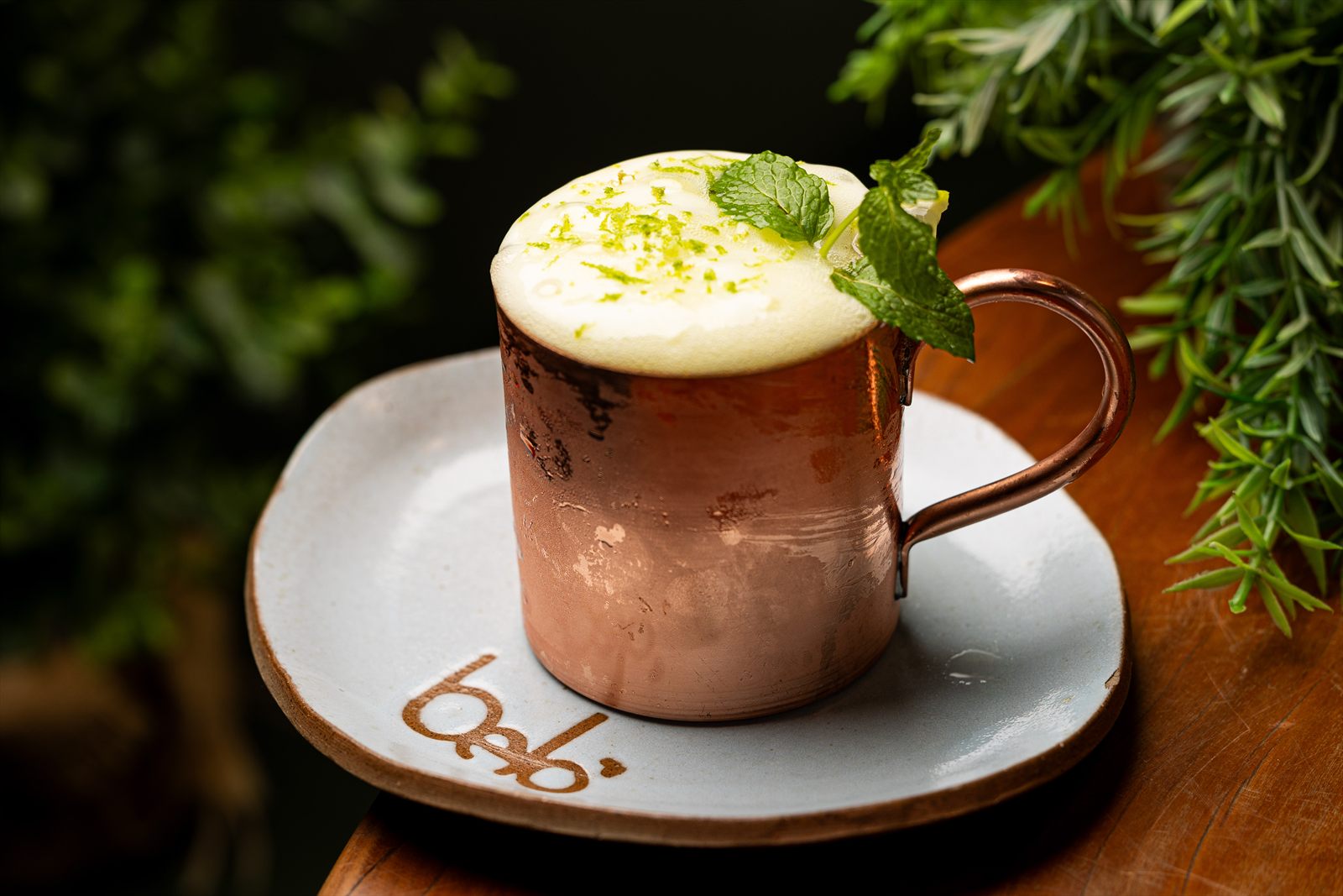 Drink em caneca de cobre coberto por espuma branca, raspas de limão e ramo de hortelã, sobre pratinho de cerâmica com selo da casa.