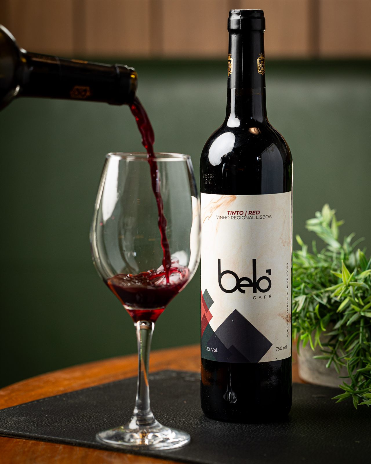 Vinho tinto Belô Café sendo servido em taça ao lado da garrafa, sobre mesa de madeira com parede verde ao fundo.